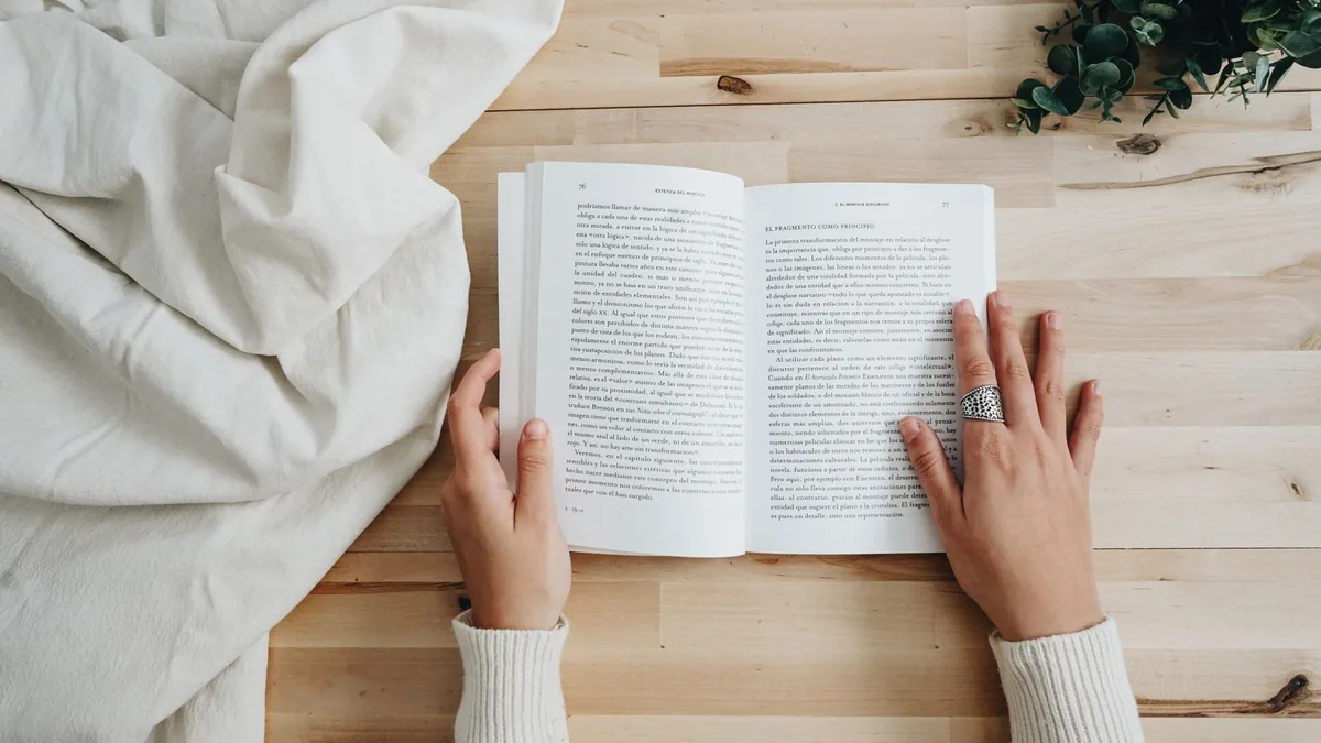 Vista superior de una persona leyendo un libro en una mesa de madera con una decoración acogedora, ideal para conceptos