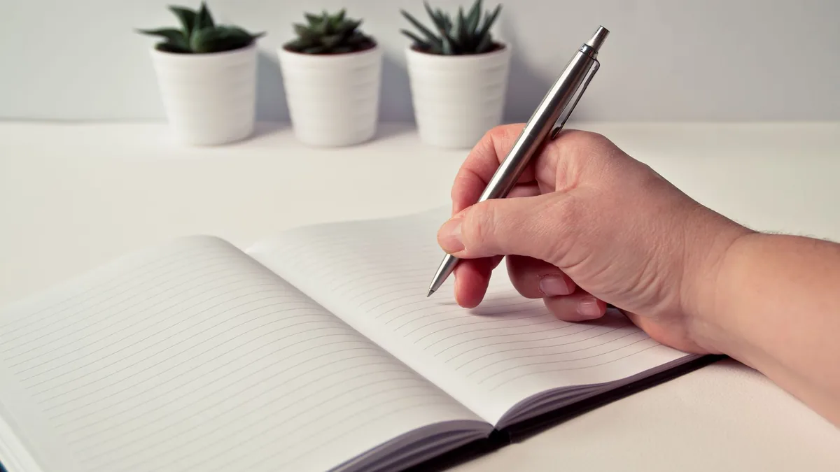 Escribiendo en un cuaderno de líneas colocado sobre una mesa con plantas suculentas al fondo.